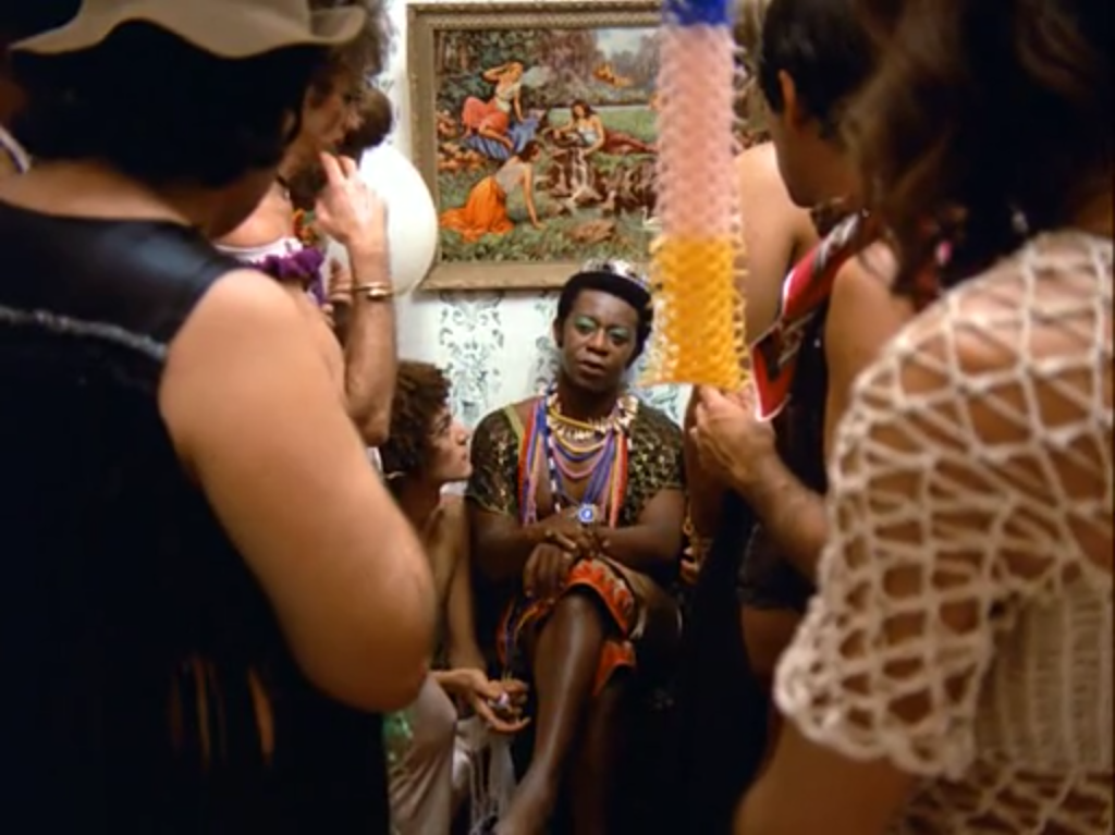 Captura de tela do filme A Rainha Diaba, com uma visão em plano de conjunto do ator Milton Gonçalves. A Rainha aparece sentada no centro da imagem, circundada pelo séquito de personagens que o filme apresenta como sua corte.
