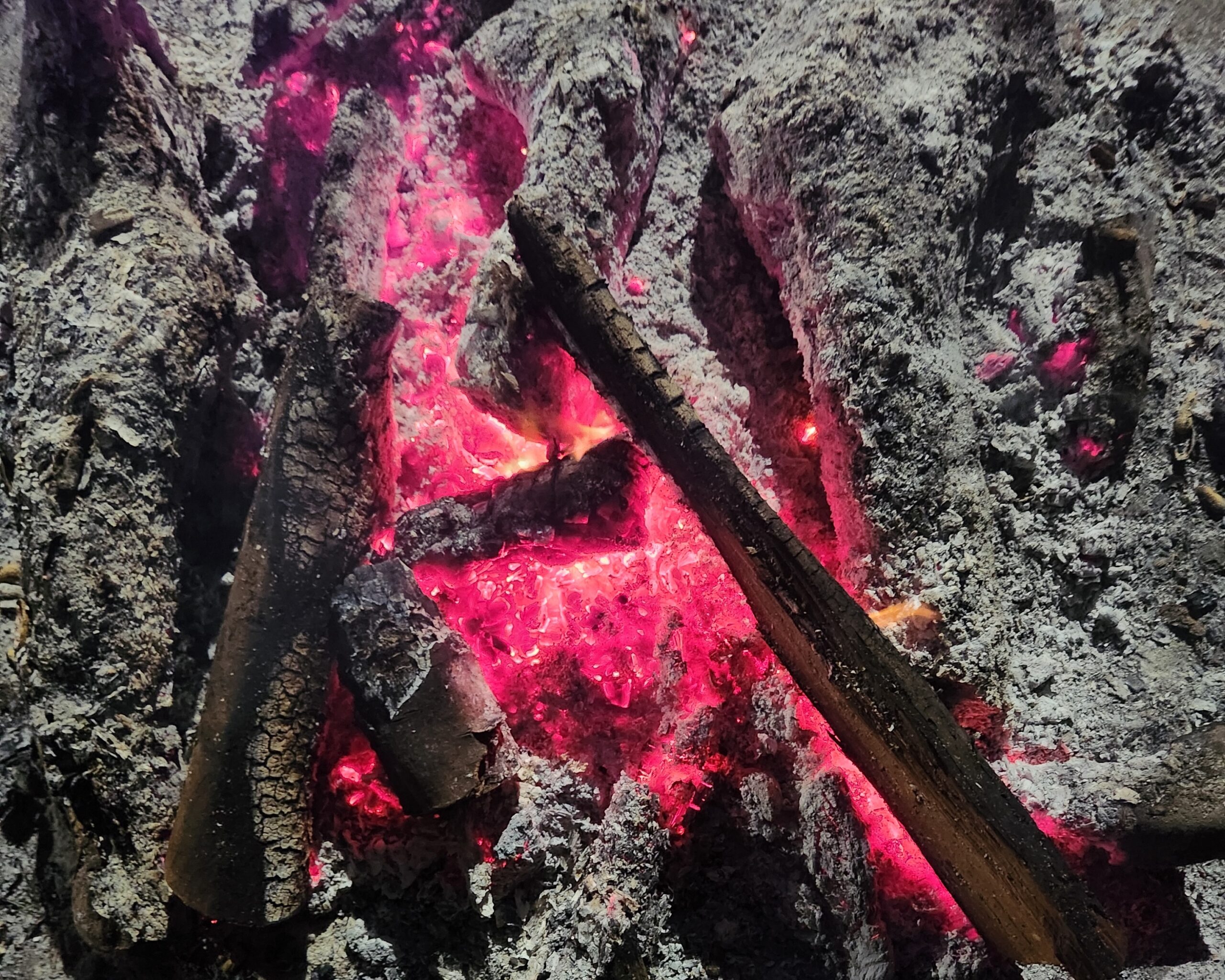 Fotografia de fogueira, vista de cima e de forma aproximada, com cinzas e alguns pedaços de madeira circundando um núcleo em que algumas chamas ainda persistem, e as cinzas em brasa aparecem em um tom vermelho e alaranjado